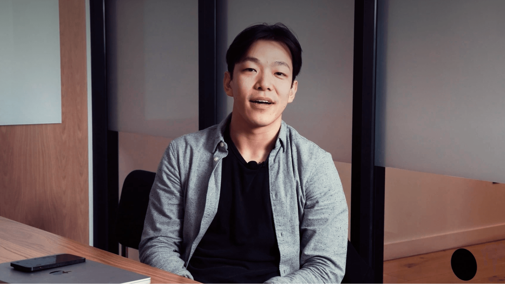 Young man seated at a desk speaking to camera, wearing gray shirt over black tee; laptop and phone nearby.